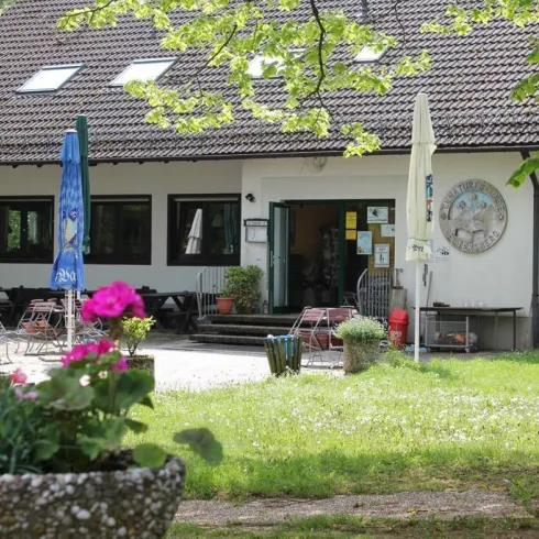 Ein gemütliches Café mit einer einladenden Terrasse und schönen Blumen. Im Hintergrund sind Bäume und eine ruhige Grünfläche zu sehen.