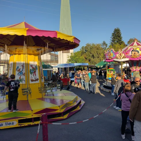 Ein Jahrmarkt mit bunten Fahrgeschäften und vielen Menschen. Die Atmosphäre ist lebhaft und fröhlich.