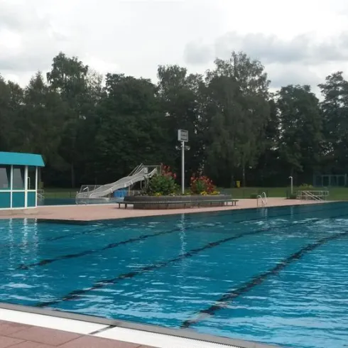 Ein Schwimmbad mit klarem Wasser und einer Rutsche. Im Hintergrund sind Bäume und eine gepflegte Umgebung zu sehen.