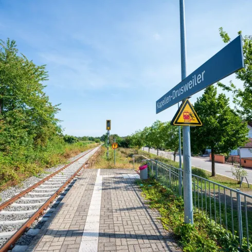 Ein ruhiger Bahnhof mit Gleisen und einer Haltestelle. Umgeben von grünen Bäumen und einem klaren blauen Himmel.
