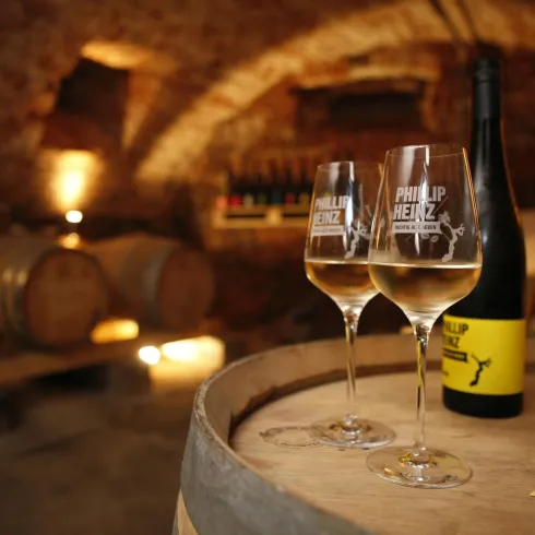 Ein gemütlicher Weinkeller mit Fässern im Hintergrund. Zwei Gläser Wein und eine Flasche stehen auf einem Holzfass.