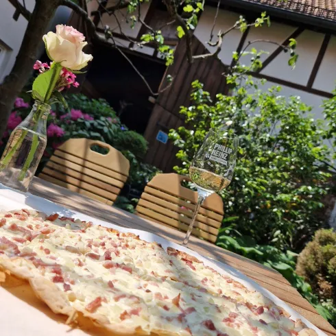 Eine frisch gebackene Flammkuchen liegt auf einem Tisch im Freien. Im Hintergrund sieht man eine grüne Umgebung mit Blumen und einem Glas Wein.