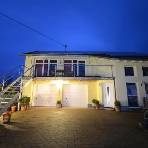 Ein modernes Haus mit einer hellen Fassade und einem gepflasterten Vorplatz. Es ist Nacht und die Fenster sind beleuchtet.