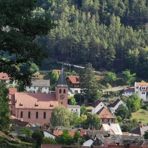 Das malerische Dorf Lindenberg (Pfalz), umgeben von Wäldern und Hügeln. Im Vordergrund steht die katholische Kirche mit ihrem charakteristischen Turm.