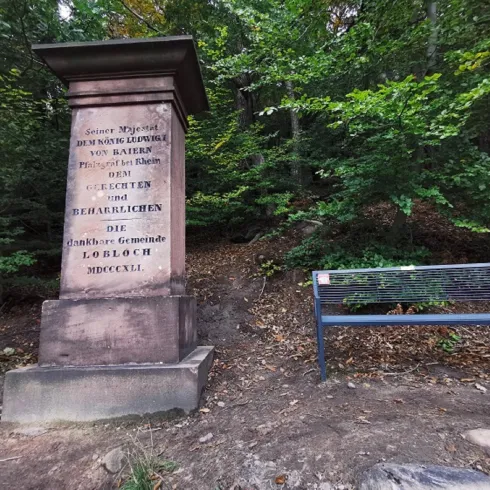 Ein Denkmal aus Stein steht im Wald, umgeben von Bäumen. Daneben befindet sich eine Holzbank zum Sitzen und Ausruhen.