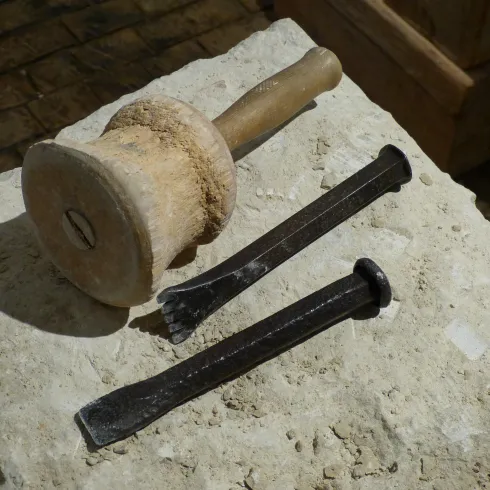Ein Steinblock mit einem Holzhammer und zwei Meißeln aus Metall liegt darauf. Die Werkzeuge sind für handwerkliche Tätigkeiten gedacht, wie zum Beispiel für den Steinmetz.
