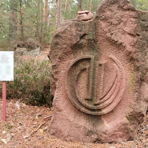 Ein großer, rotbrauner Stein mit einer eingravierten Symbolik steht in einem Wald. Neben dem Stein befindet sich ein Informationsschild.