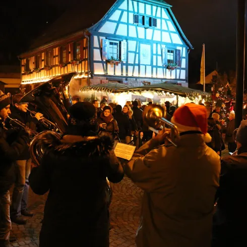 Eine festliche Szene mit einer Menschenmenge, die einem Weihnachtsmarkt bei Nacht lauscht. Lichter und eine festliche Atmosphäre umgeben das traditionelle Fachwerkhaus.