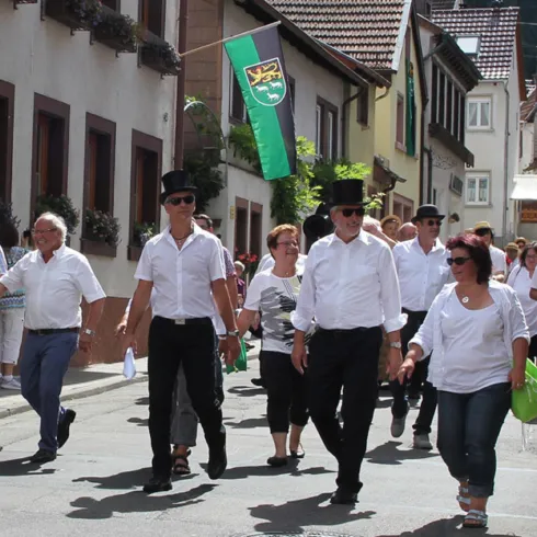 Eine Gruppe von Menschen in traditioneller Kleidung geht eine Straße entlang. Im Hintergrund sind Häuser und eine Fahne sichtbar.