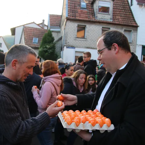 Eine Menschenmenge auf der Straße, die an einem Fest teilnimmt. Zwei Männer tauschen orangefarbene Eier aus.