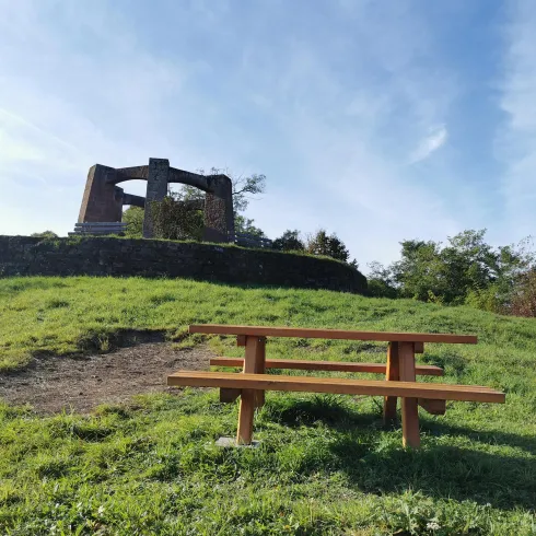 Ein Holzpicknicktisch mit Bänken steht auf einer grünen Wiese. Im Hintergrund das Kriegerdenkmal von Weidenthal und ein blauer Himmel zu sehen.