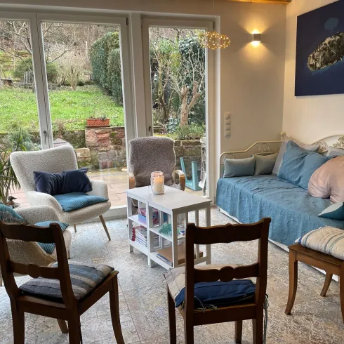 Ein helles Wohnzimmer mit bequemen Sofas und Stühlen. Große Fenster bieten einen Blick auf den Garten. Dieser Raum wird für Trauergruppen genutzt.