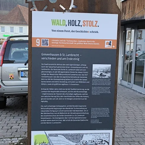 Ein informatives Schild über den Wald, Holz und die lokale Geschichte. Es enthält Texte und Bilder zu diesen Themen und ist in einer städtischen Umgebung platziert.