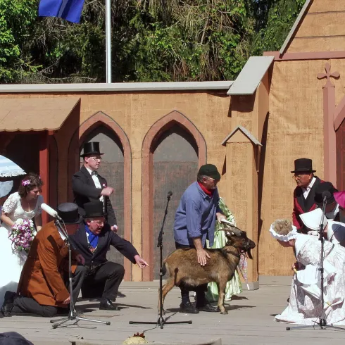 Eine Theateraufführung mit Menschen in historischen Kostümen. In der Mitte der Bühne steht eine Ziege, während die Darsteller interagieren.