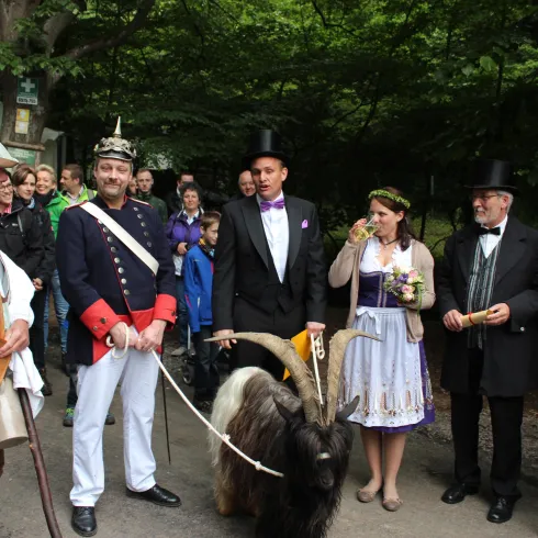 Eine Gruppe von Menschen in traditioneller Kleidung steht im Freien. Ein Ziegenbock ist ebenfalls anwesend, während die Leute lächeln und miteinander interagieren.