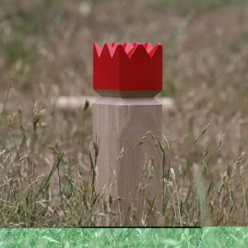 Ein Holzpfeiler mit einer roten Krone steht im Gras. Die Krone hat eine gezackte Oberkante. Dies ist ein Spielelement vom Wikingerschach.