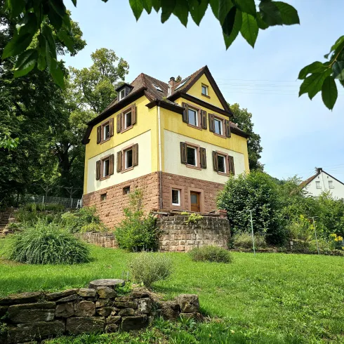 Ein charmantes, gelbes Haus mit einem grünen Garten und vielen Bäumen. Der blauen Himmel und die natürliche Umgebung schaffen eine ruhige Atmosphäre.