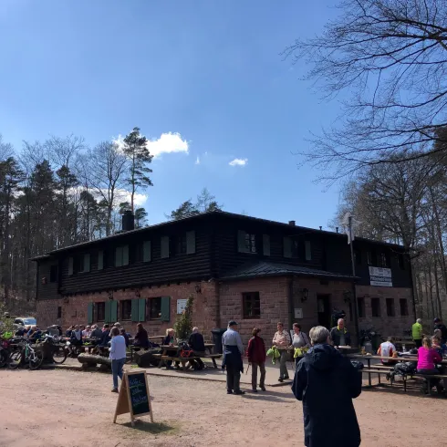 Ein gemütliches Restaurant im Wald mit vielen Gästen. Draußen stehen Bänke und Tische, und die Sonne scheint.