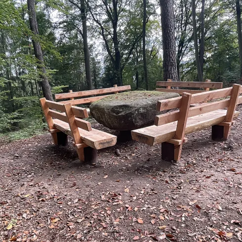 Eine rustikale Sitzecke aus Holz mit einem großen Stein in der Mitte. Umgeben von Bäumen im Wald.