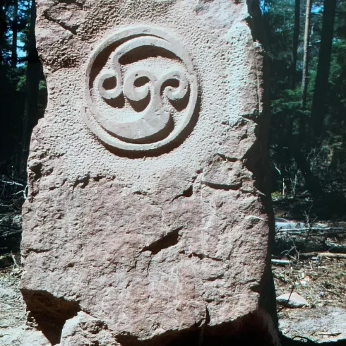 Ein großer, aufrechter Stein mit einer kunstvollen Gravur in der Mitte. Der Stein steht in einem bewaldeten Bereich.
