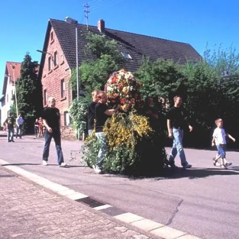 Eine festliche Parade mit Blumenkränzen auf der Straße. Menschen gehen zusammen und feiern in einer ländlichen Umgebung.