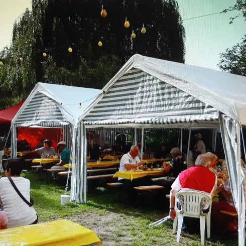 Ein gemütliches Gartenfest unter Zelten. Viele Menschen sitzen an langen Tischen und genießen die Atmosphäre.