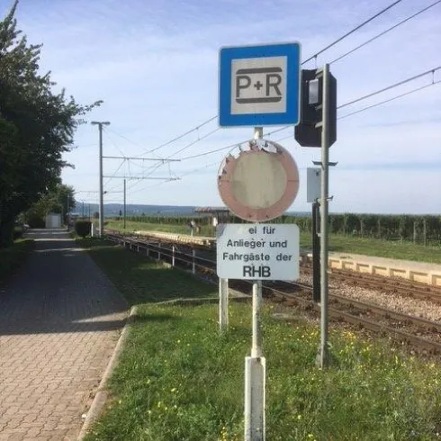 Ein Parkplatz für Fahrgäste der RHB ist in der Nähe des Bahnhofs gekennzeichnet. Der Weg ist von Gras und Bäumen umgeben.