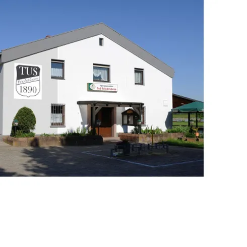 Ein weißes Gebäude mit der Aufschrift „TUS 1890“ an der Fassade. Es gibt einen kleinen Gartenbereich mit Tischen und Stühlen.