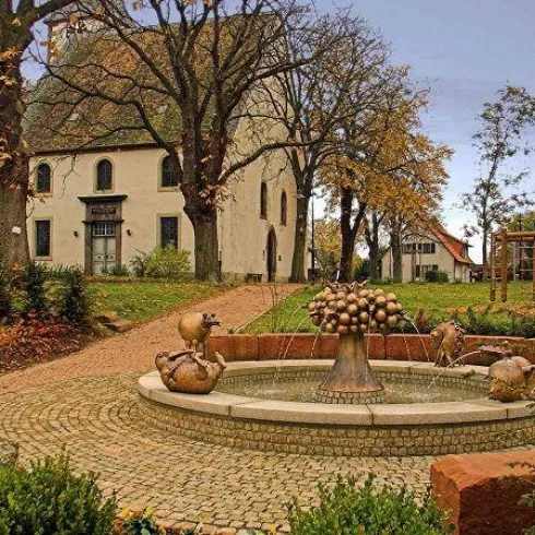 Ein schöner Platz mit einem Brunnen und einer historischen Gebäude im Hintergrund. Bäume und Sträucher umgeben die Szene in einer ruhigen Umgebung.