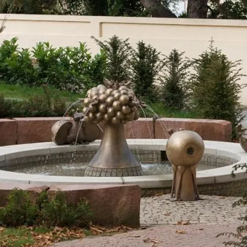 Ein moderner Brunnen mit stilisierten, bronzeartigen Skulpturen in einem Garten. Umgeben von grünen Pflanzen und Steinmauern.