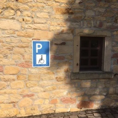 Ein Parkplatzschild für Personen mit Behinderung an einer Steingemauer. Im Hintergrund ist ein kleines Fenster zu sehen.