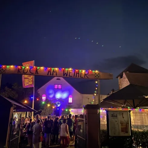 Eine lebendige Veranstaltung mit vielen Menschen unter dem Schild „Rock am Wehner“. Bunte Lichter erleuchten die Umgebung bei Dämmerung.