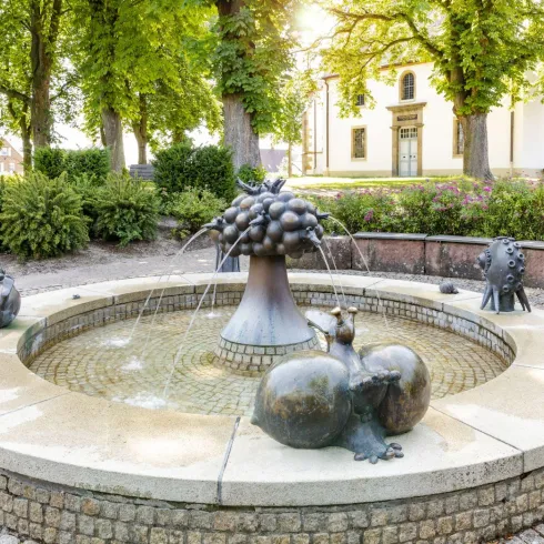 Ein Brunnen mit künstlerischen Skulpturen von Früchten und Tieren. Umgeben von Bäumen und einer grünen Landschaft.