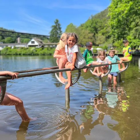 Kinder spielen am Ufer eines Gewässers und sitzen auf einem Handlauf. Einige halten ihre Füße im Wasser, während andere fröhlich lächeln.