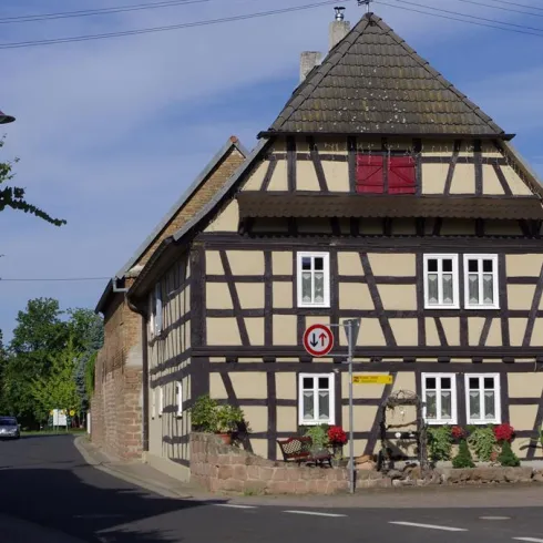 Ein traditionelles Fachwerkhaus an einer Straßenecke mit blauen Himmel im Hintergrund. Die Fassade ist hellbraun mit weißen Fenstern und roten Akzenten.