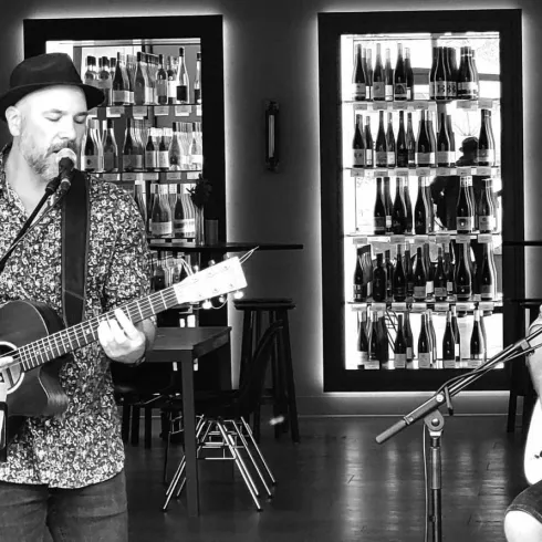 Zwei Musiker spielen live in einem stilvollen Raum. Im Hintergrund sind Regale mit verschiedenen Getränken zu sehen.