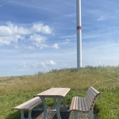 Eine Sitzbank mit Tisch steht auf einer Wiese. Im Hintergrund ist ein Windrad und blauer Himmel zu sehen.