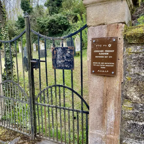 Ein rustikales Tor zu einem historischen jüdischen Friedhof, umgeben von grüner Natur. An der Mauer befindet sich ein informatives Schild.