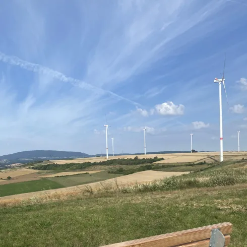 Eine Holzbank steht auf einer Wiese mit Blick auf grüne Felder und Windkraftanlagen. Der Himmel ist blau mit einigen Wolken.