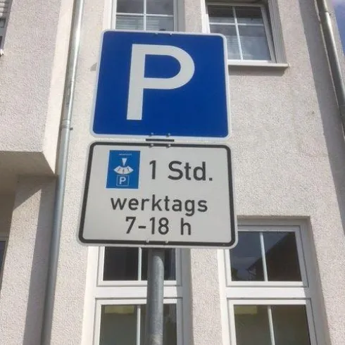 Ein Parkplatzschild mit dem Symbol „P“ in Blau. Es zeigt, dass das Parken an Werktagen von 7 bis 18 Uhr für 1 Stunde erlaubt ist.