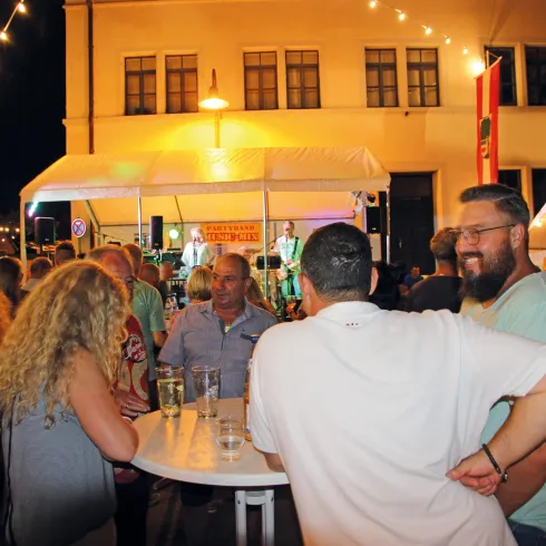 Eine lebhafte Veranstaltung bei Nacht mit vielen Menschen, die miteinander sprechen und Spaß haben. Im Hintergrund spielt eine Band, und die Atmosphäre ist festlich.
