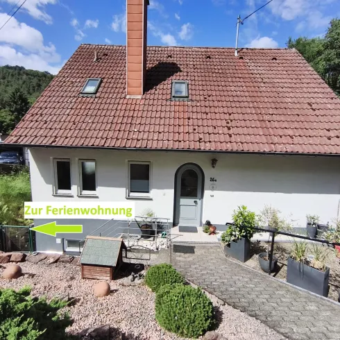 Ein kleines, einladendes Haus mit rotem Dach und einem gepflegten Vorgarten. Ein Weg führt zur Ferienwohnung, die deutlich gekennzeichnet ist.