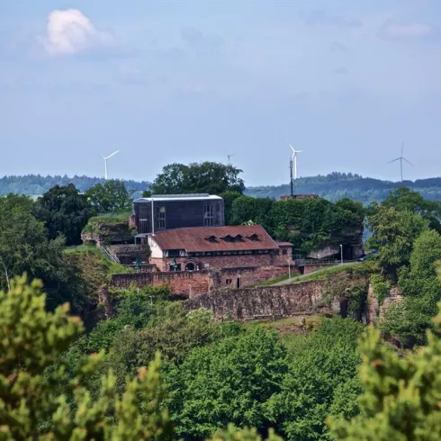 Eine alte Festung auf einem Hügel, umgeben von Bäumen und grüner Landschaft. Im Hintergrund sind Windräder sichtbar.