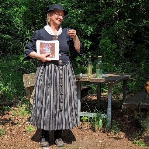Eine Frau in traditioneller Kleidung steht in einer grünen Umgebung und hält ein Buch in der Hand. Auf einem Tisch neben ihr stehen zwei Flaschen und weitere Gegenstände.
