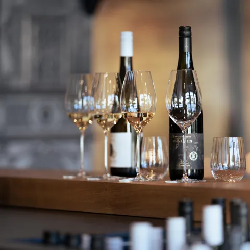 Eine stilvolle Anordnung von Weingläsern und Flaschen auf einer Holzoberfläche. Im Hintergrund sind weitere dekorative Elemente zu sehen.