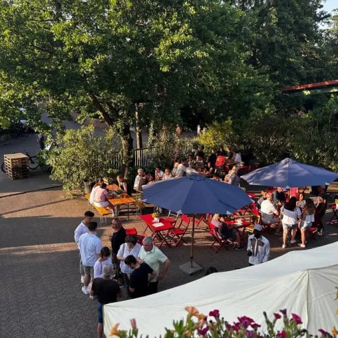 Eine belebte Außenbereich mit mehreren Tischen und Sonnenschirmen. Menschen genießen die Zeit im Freien unter einem großen Baum.