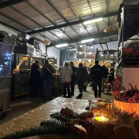 Ein lebhafter Innenraum mit vielen Menschen, die in einem Lager stehen. Auf einem Tisch im Vordergrund sind Kerzen und dekoratives Grün zu sehen.