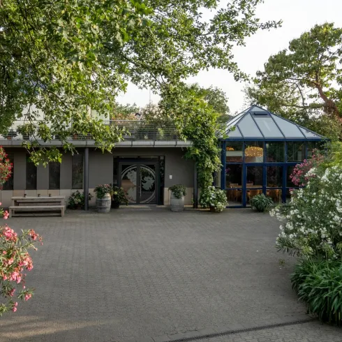 Ein einladender Eingangsbereich mit bunten Blumen und grünen Pflanzen. Im Hintergrund ist ein modernes Gebäude mit gläsernem Dach zu sehen.