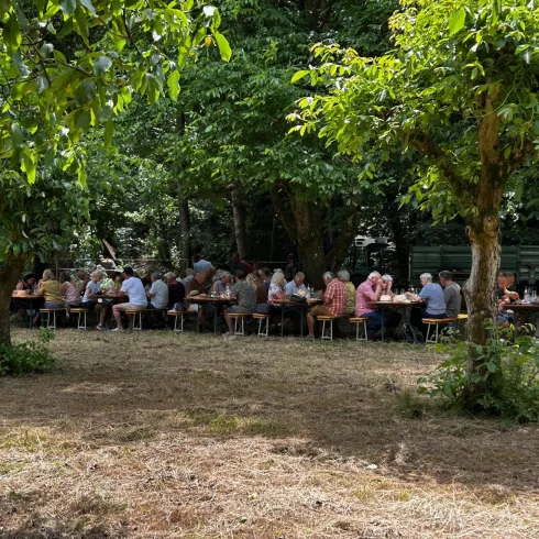 Eine Gruppe von Menschen sitzt an langen Tischen im Freien unter Bäumen. Die Atmosphäre ist entspannt und freundlich.