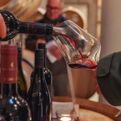 Ein Weinglas wird mit roten Wein gefüllt. Im Hintergrund sind unscharfe Personen und Flaschen zu sehen.